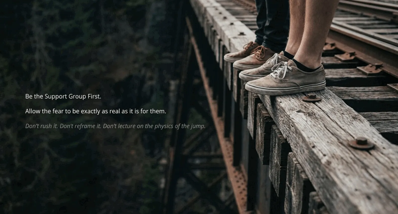 Close-up of two pairs of shoes standing on the edge of a wooden railroad trestle high above a forested drop, with the words "Be the Support Group First. Allow the fear to be exactly as real as it is for them. Don't rush it. Don't reframe it. Don't lecture on the physics of the jump." overlaid in the lower left.